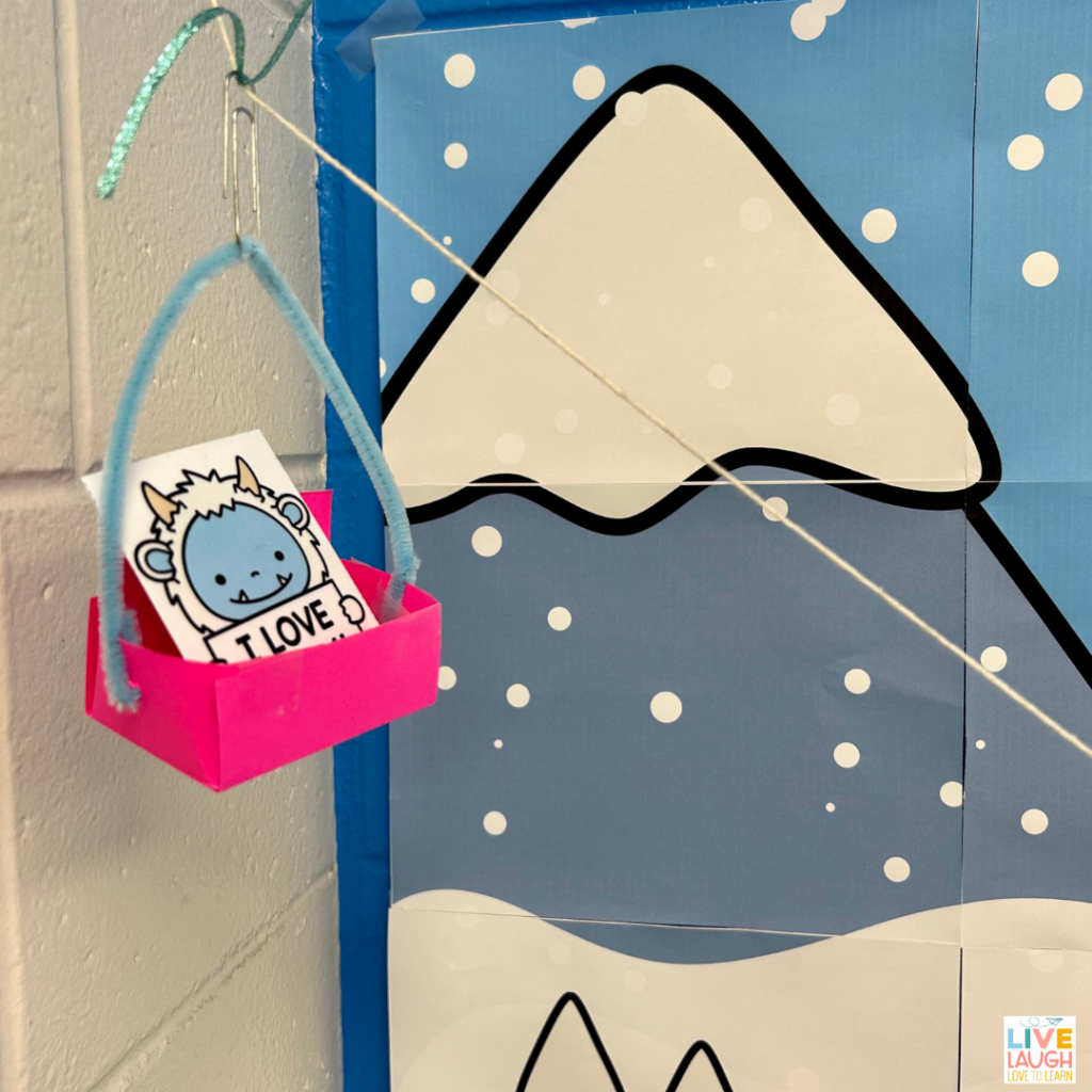 photo of a paper yeti in a pink paper holder with a blue pipe cleaner holding it on to a paper clip.  The paper holder is at the top of a string that is angled downward. In the background is a poster of a drawing of a snow covered mountain.