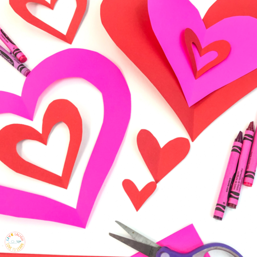 Image of red and pink cut out Valentine's Day paper hearts with pink and red crayons and a pair of scissors.