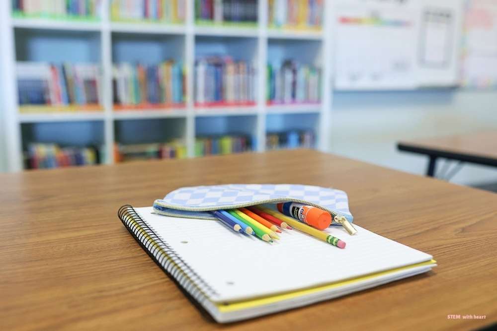 A neatly organized spiral notebook and colorful pencil pouch on a classroom desk, with a tidy classroom bookshelf in the background — simple end-of-year classroom organization tips for elementary teachers.