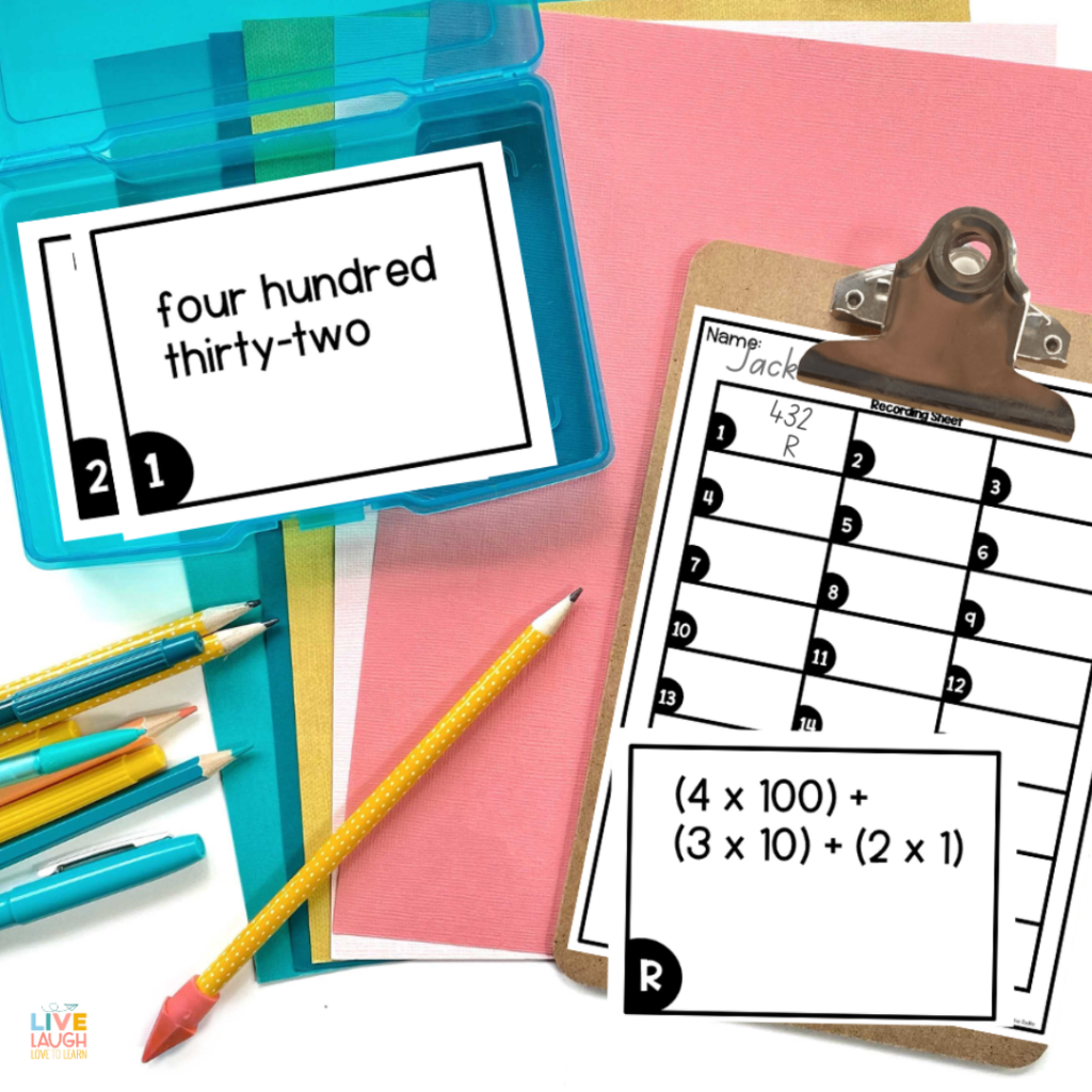 An image shows a place value task card in a photo box that reads "four hundred thirty-two". Next to the photo box is a clipboard with a task card recording page. On the clipboard is a task card that reads "(4x100)+(3x10)+(2x1). There is also a set of pens and pencils laying beside the clipboard.