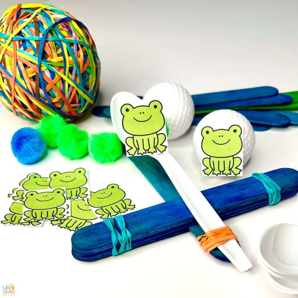 an image of catapult made from craft sticks and a plastic spoon. A printed paper green smiling frog is attached to a pom pom and sitting on the spoon.
