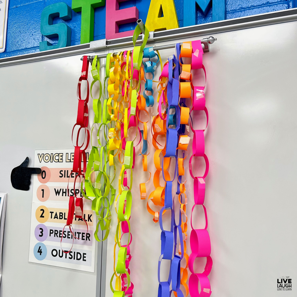 The image displays a classroom whiteboard with colorful paper chains hanging from a rod, organized by different bright colors such as red, yellow, green, pink, orange, and blue. Above the paper chains, large, colorful letters spelling "STEAM" are visible. The setting suggests a vibrant and engaging STEM classroom environment.