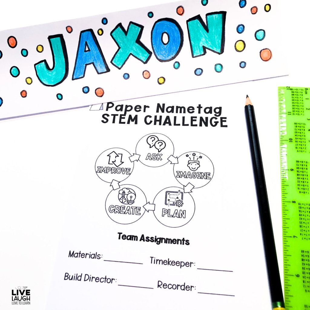The image shows materials for a "Paper Nametag STEM Challenge." At the top, there is a brightly colored name tag with the name "Jaxon" written in large blue and green bubble letters, surrounded by colorful dots. Below it is a worksheet titled "Paper Nametag STEM Challenge" that outlines the engineering design process with steps: Ask, Imagine, Plan, Create, and Improve. The sheet also includes team assignments for roles like Timekeeper, Build Director, and Recorder. A pencil and a green ruler are placed next to the worksheet. The setting is creative and organized for a classroom activity.