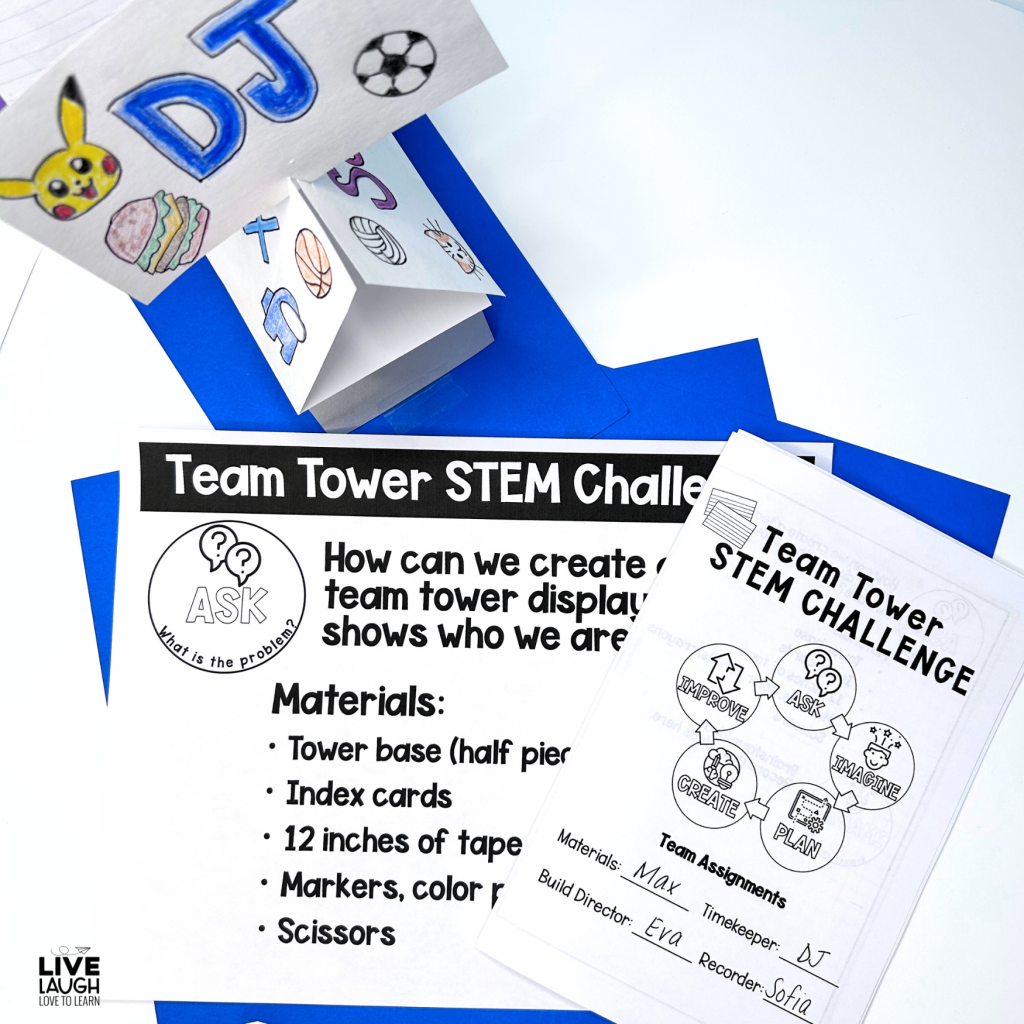 An image of materials for a 'Team Tower STEM Challenge' in a classroom setting. The challenge sheet includes instructions, a list of materials needed (such as index cards, tape, markers, and scissors), and a section for team assignments. The challenge focuses on creating a team tower display that reflects the team's identity. A folded paper tower with drawings of various symbols and a student's name 'DJ' is visible. The image is branded with a 'Live Laugh Love to Learn' logo in the corner