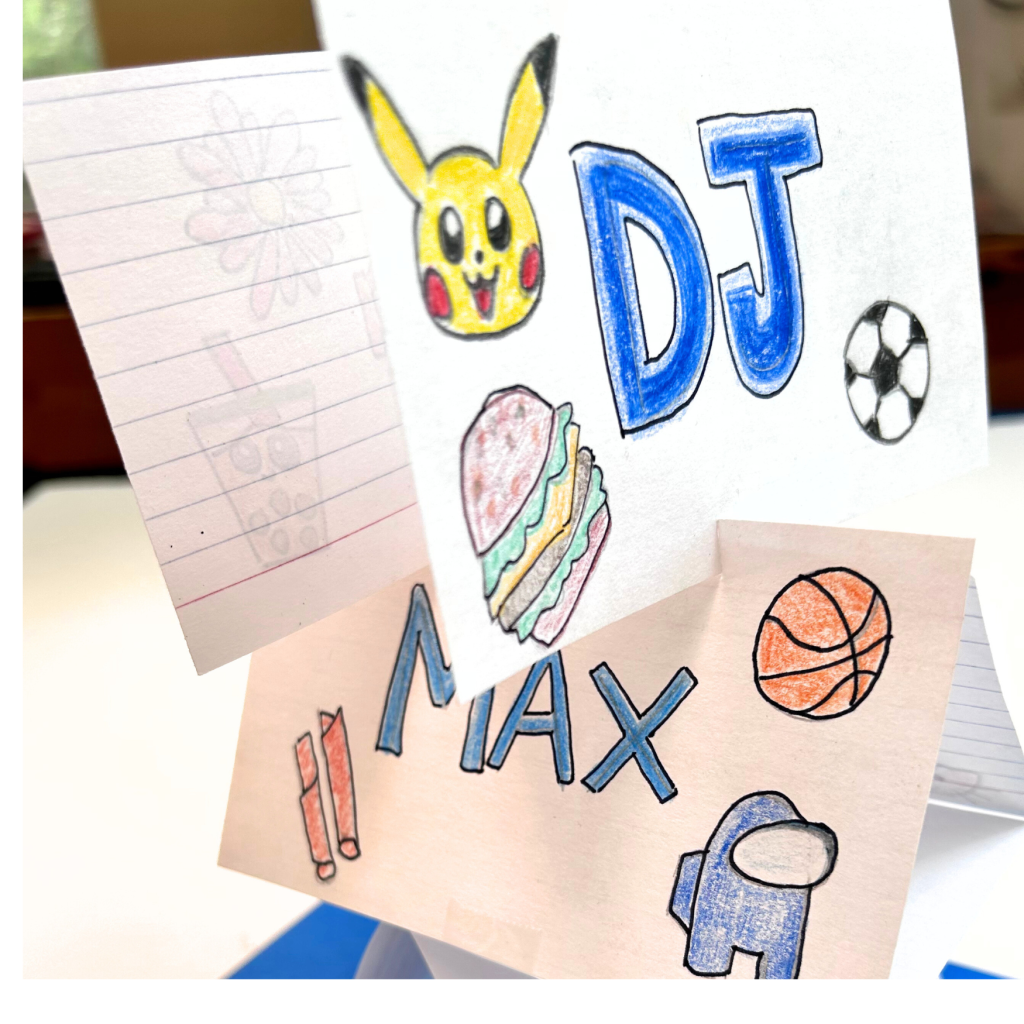 A close-up of a folded paper tower with colorful drawings and names created by students for a STEM challenge. The name 'DJ' is prominently displayed alongside drawings of a Pikachu, a soccer ball, and a burger. Below it, the name 'Max' is shown with illustrations of a basketball, a pair of Takis, and a character resembling the Among Us game. The paper cards are part of a team identity activity in the 'Team Tower STEM Challenge.