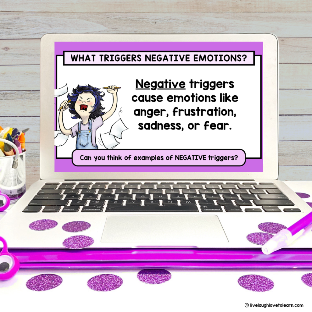 laptop displaying a digital resource titled "What Triggers Negative Emotions," with an illustration of a girl throwing paper in the air with a look of frustration next to a definition of "Negative triggers." The laptop is on a wooden desk with purple glittery dots, a purple folder, and classroom supplies like markers and scissors nearby.