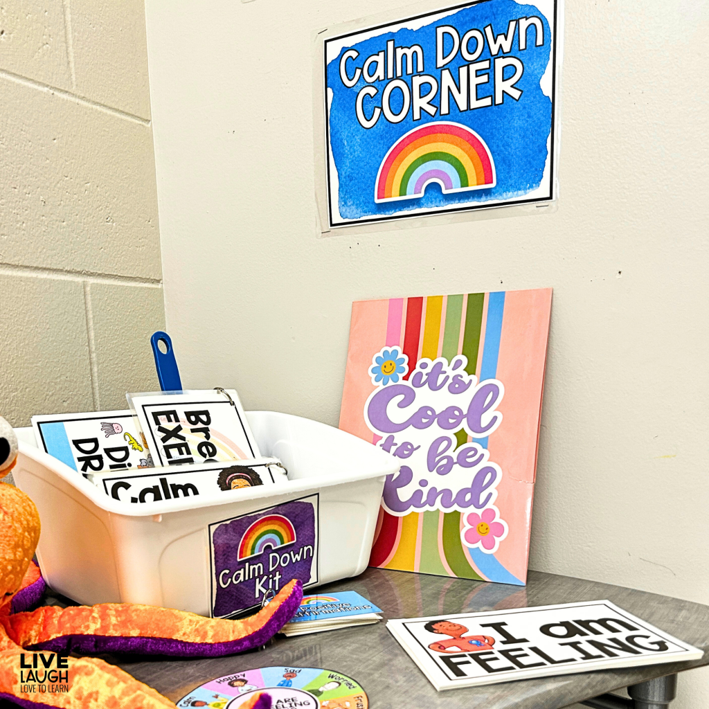 A classroom Calm Down Corner setup featuring a blue sign with 'Calm Down Corner' and a rainbow illustration on the wall. A white bin labeled 'Calm Down Kit' contains laminated breathing exercises and calm-down strategy cards. A colorful sign reading 'It's Cool to be Kind' leans against the wall. A plush octopus, an emotions wheel, and an 'I am FEELING' check-in card are also present on the table, creating a welcoming and supportive space for emotional regulation.