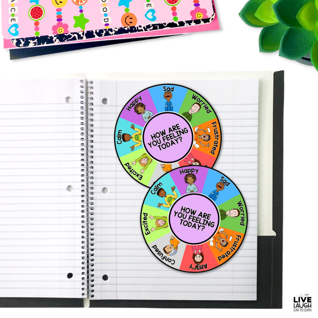 A spiral notebook lies open on a desk with two colorful emotion wheels placed on top. Each wheel features a variety of emotions, such as happy, sad, worried, frustrated, excited, and calm, represented by diverse illustrated children. The center of each wheel asks, "How are you feeling today?" A pink and black composition notebook and a decorated folder with smiley faces, stars, and beads are partially visible in the background. A small green succulent plant sits in the top right corner. The image includes a "Live Laugh Love to Learn" watermark in the bottom right corner.