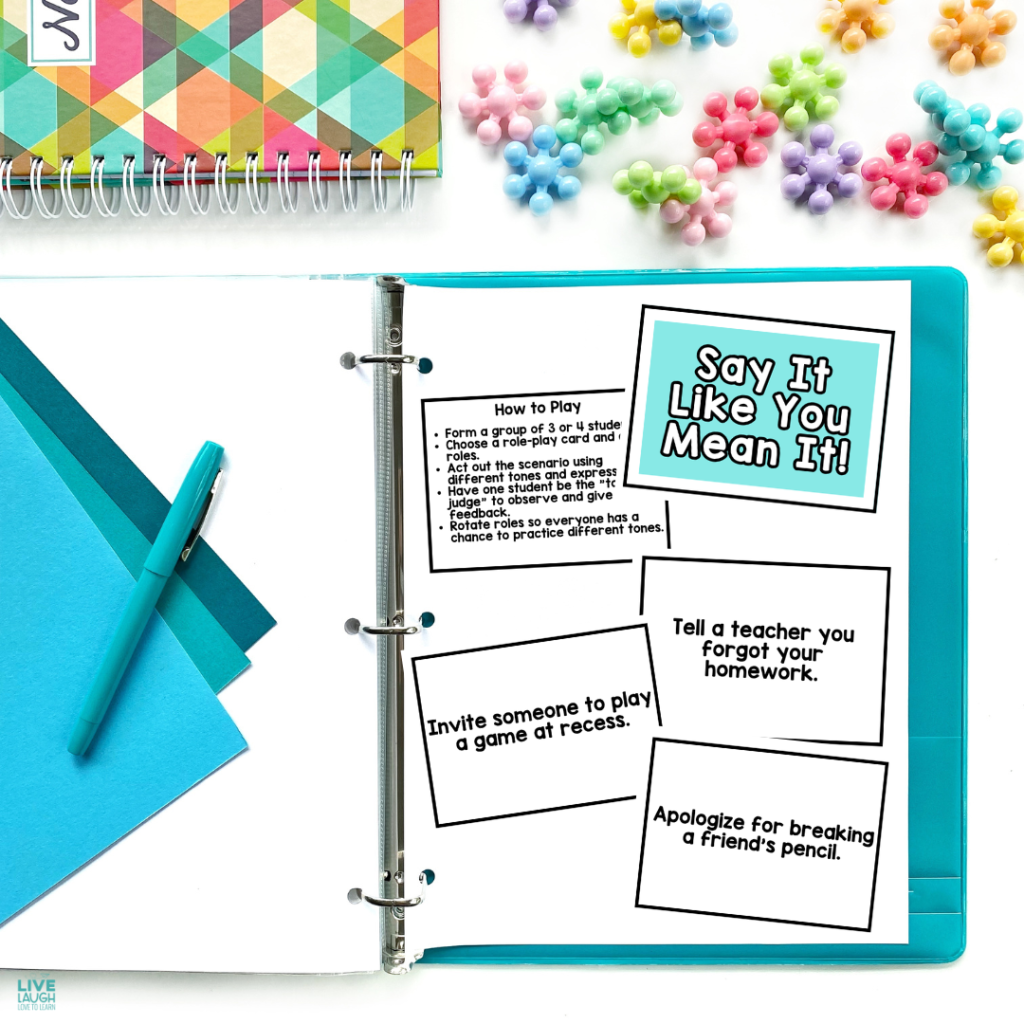 An open binder displays the "Say It Like You Mean It!" SEL communication activity. It includes instructions for students to role-play everyday scenarios using different tones, with prompts like “Tell a teacher you forgot your homework,” “Invite someone to play a game at recess,” and “Apologize for breaking a friend’s pencil.” A teal pen, pastel push pins, and colorful paper surround the binder, creating a bright, organized classroom setting.