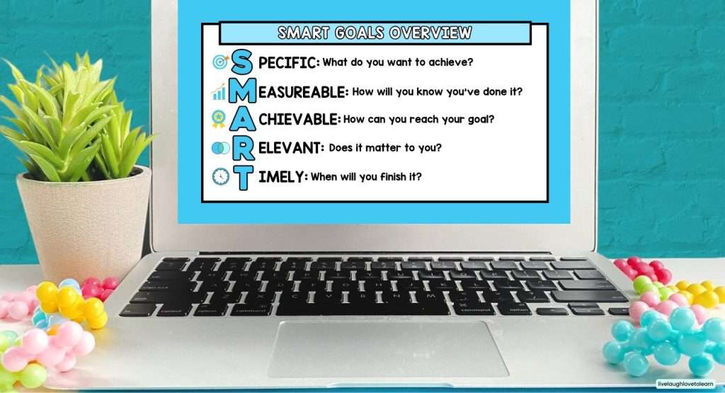 Laptop displaying a SMART Goals Overview slide with a blue border. The screen defines each letter of SMART: Specific (What do you want to achieve?), Measurable (How will you know you’ve done it?), Achievable (How can you reach your goal?), Relevant (Does it matter to you?), and Timely (When will you finish it?). The laptop sits on a bright blue surface with colorful beads and a small green plant beside it.