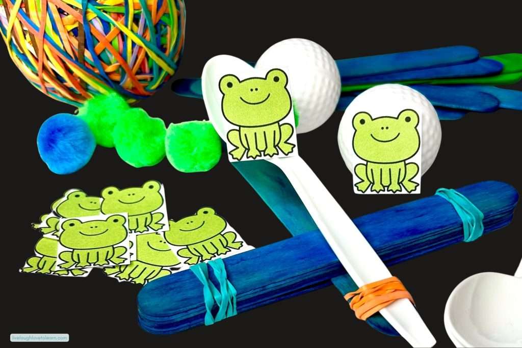 End of year STEM activity of a popsicle stick catapult held together with rubber bands, with a plastic spoon arm and a frog illustration on the spoon, surrounded by golf balls, pom poms, and extra craft sticks on a black background.