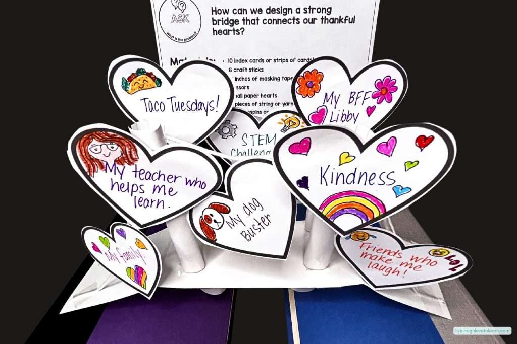 A student-made bridge built from rolled paper tubes and cardstock, topped with heart-shaped cutouts decorated with colorful drawings and phrases like "My teacher who helps me learn," "Kindness," and "Friends who make me laugh."