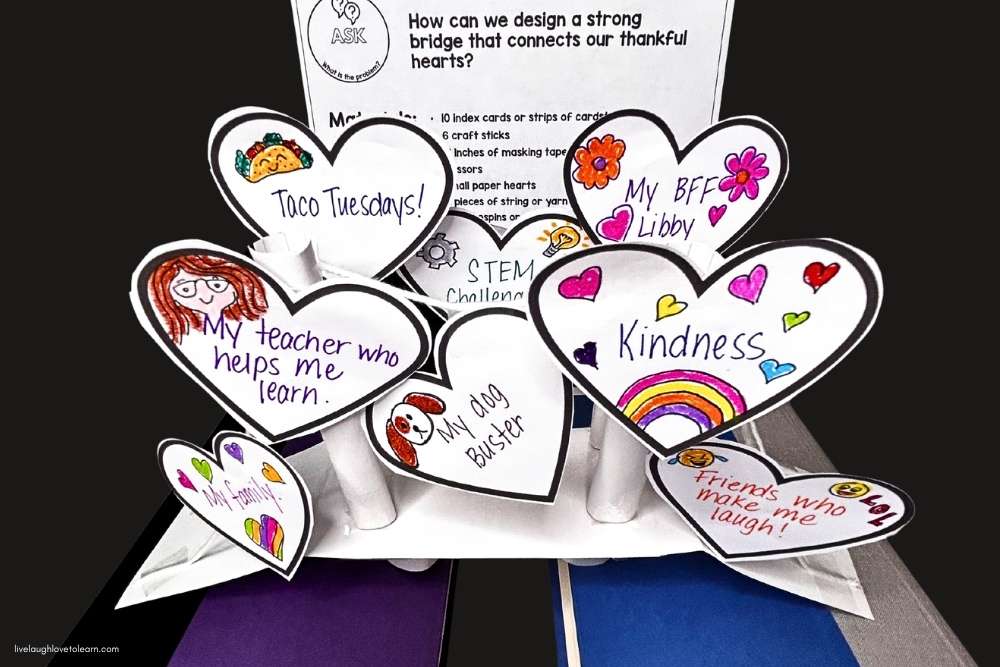A student-made bridge built from rolled paper tubes and cardstock, topped with heart-shaped cutouts decorated with colorful drawings and phrases like "My teacher who helps me learn," "Kindness," and "Friends who make me laugh."