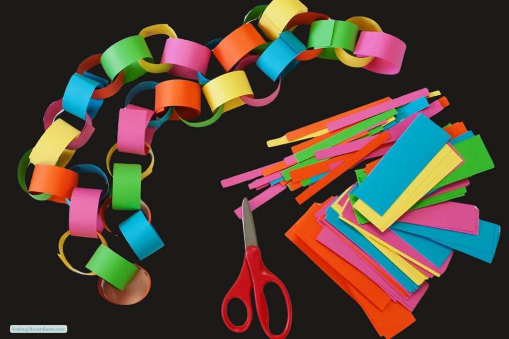 A long colorful paper chain made from bright pink, orange, yellow, green, and blue paper loops, displayed on a black background next to red scissors and a pile of pre-cut paper strips for an end of year STEM activity.