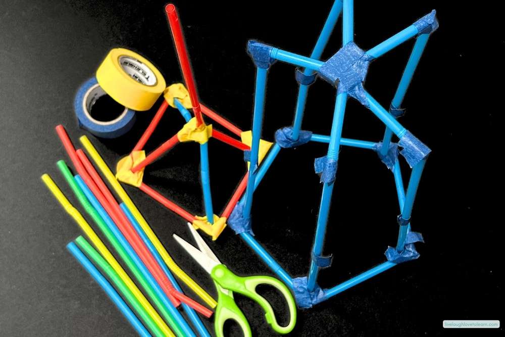 Two straw-and-tape towers in different geometric designs, one small yellow structure and one larger blue cube-like frame,  displayed alongside colorful straws, green scissors, and rolls of masking tape on a black background for an end of year STEM challenge.
